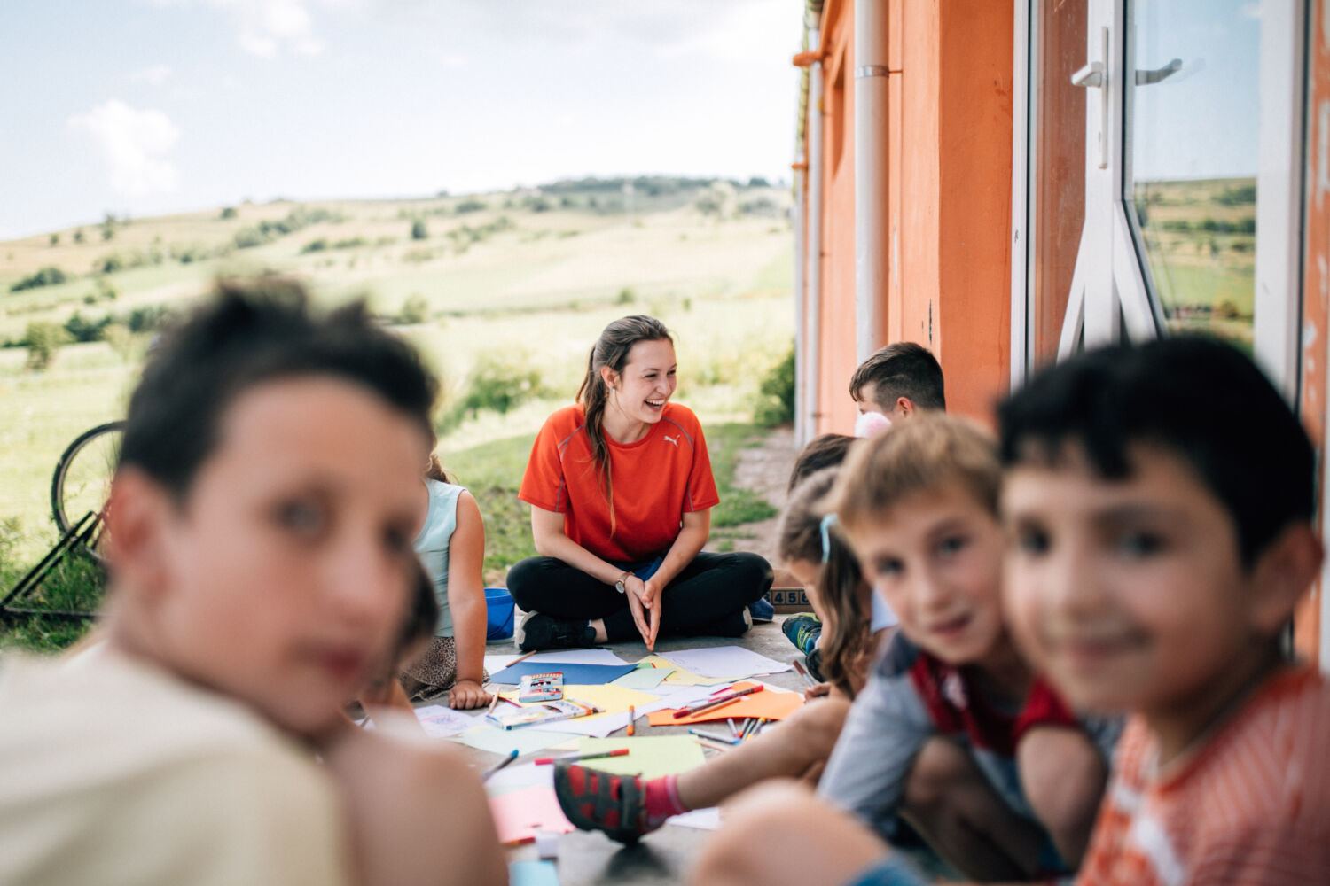 Een vrijwilliger zit met kinderen aan het knutselmateriaal onder een open afdak, met een weiland erachter