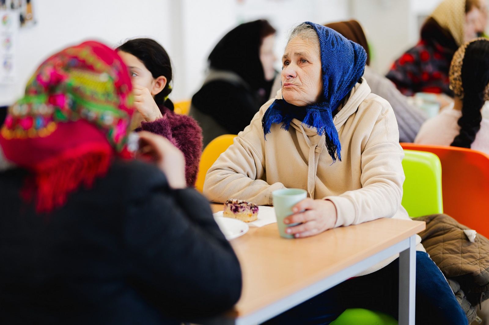 Een oudere vrouw met een blauwe hoofddoek bij een kop thee en een stuk taart